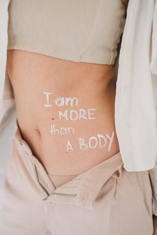 Image shows a person standing with only their torso in frame, with the words "I am more than a body" written on their skin in white ink.