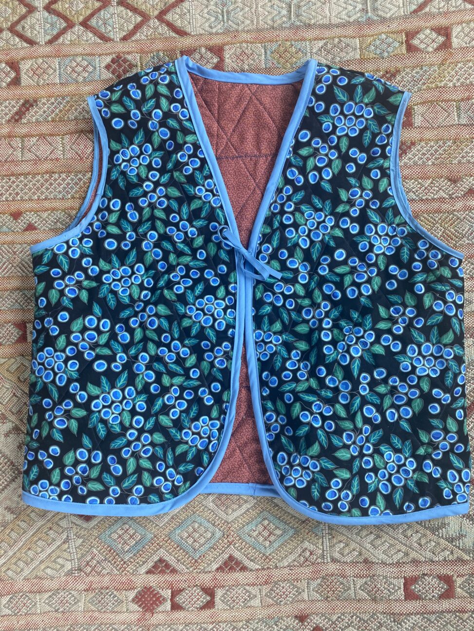 Photo shows a vest made by Alana Norie. The outside fabric is black cotton, with blueberries and leafs in a pattern on top. The vest is lined with a moody pink and quilted in a diamond pattern. The edges of the vest have a light blue bias and there is a blue bow on the front.